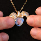 A Collar de Corazón Cofre personalizado con alas de ángel y gemas pequeñas muestra la foto recuerdo de una mujer mayor. Dos dedos sostienen el collar sobre una tela negra.