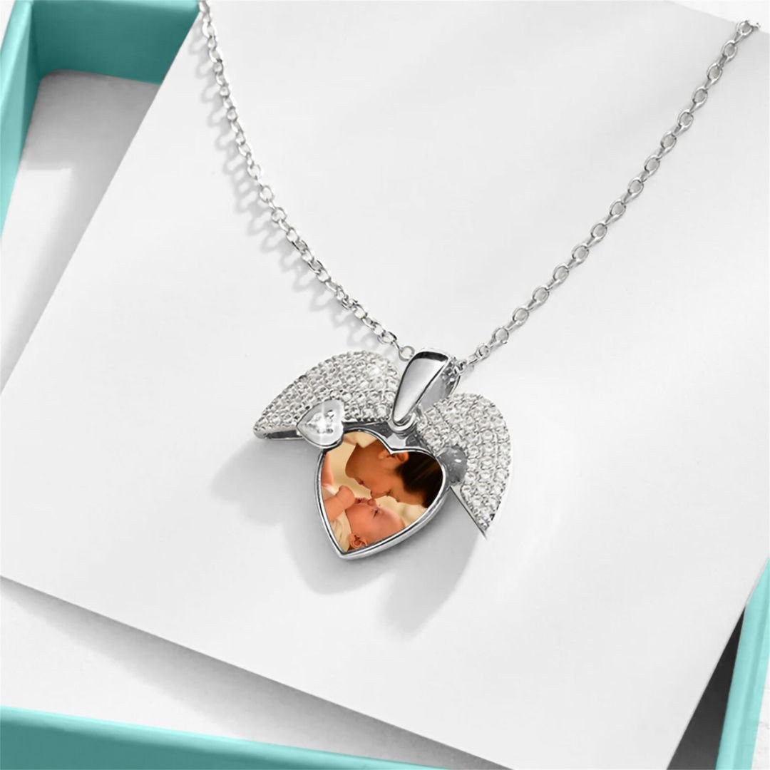 El Collar de Corazón Cofre personalizado es un collar de corazón de plata con alas de ángel, una pequeña gema y pétalos de melocotón preservados en su centro transparente, presentado en un joyero azul sobre fondo blanco.