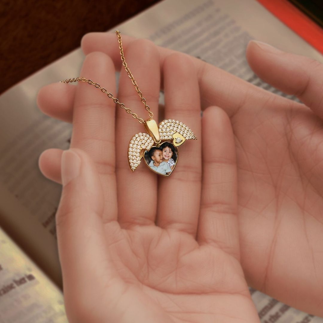 Una mano sostiene un Collar de Corazón Cofre personalizado en color dorado, decorado con alas de ángel y un foto recuerdo de dos niños sonriendo en su interior, mientras un libro abierto aparece al fondo.