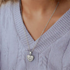 El Collar de Corazón Cofre personalizado, bañado en plata y con incrustaciones de gemas, cuelga delicadamente del cuello de una persona que luce un suéter tejido lavanda claro.
