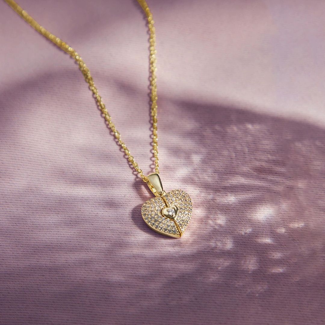 El Collar de Corazón Cofre personalizado presenta un colgante en forma de corazón con piedras brillantes y un motivo de ojo de cerradura, elegantemente engastado sobre un fondo de suave textura rosa claro.