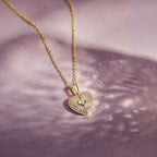 El Collar de Corazón Cofre personalizado presenta un colgante en forma de corazón con piedras brillantes y un motivo de ojo de cerradura, elegantemente engastado sobre un fondo de suave textura rosa claro.