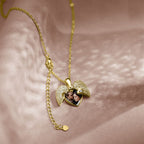 El Collar de Corazón Cofre personalizado, un medallón de corazón dorado con alas y piedras brillantes, descansa sobre una suave tela rosa. Al abrirlo, revela una pequeña foto recuerdo de dos personas sonrientes en su interior.