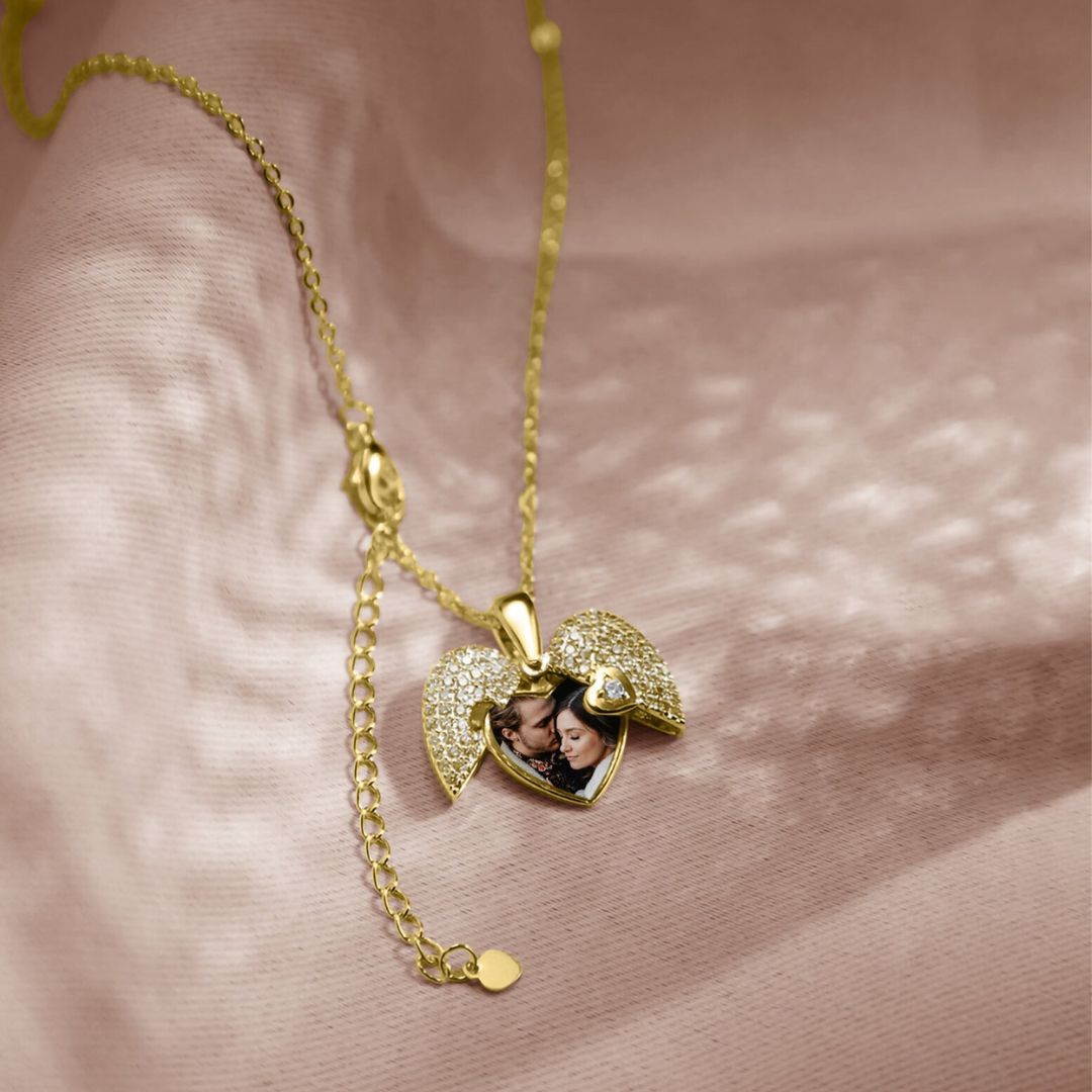El Collar de Corazón Cofre personalizado, un medallón de corazón dorado con alas y piedras brillantes, descansa sobre una suave tela rosa. Al abrirlo, revela una pequeña foto recuerdo de dos personas sonrientes en su interior.