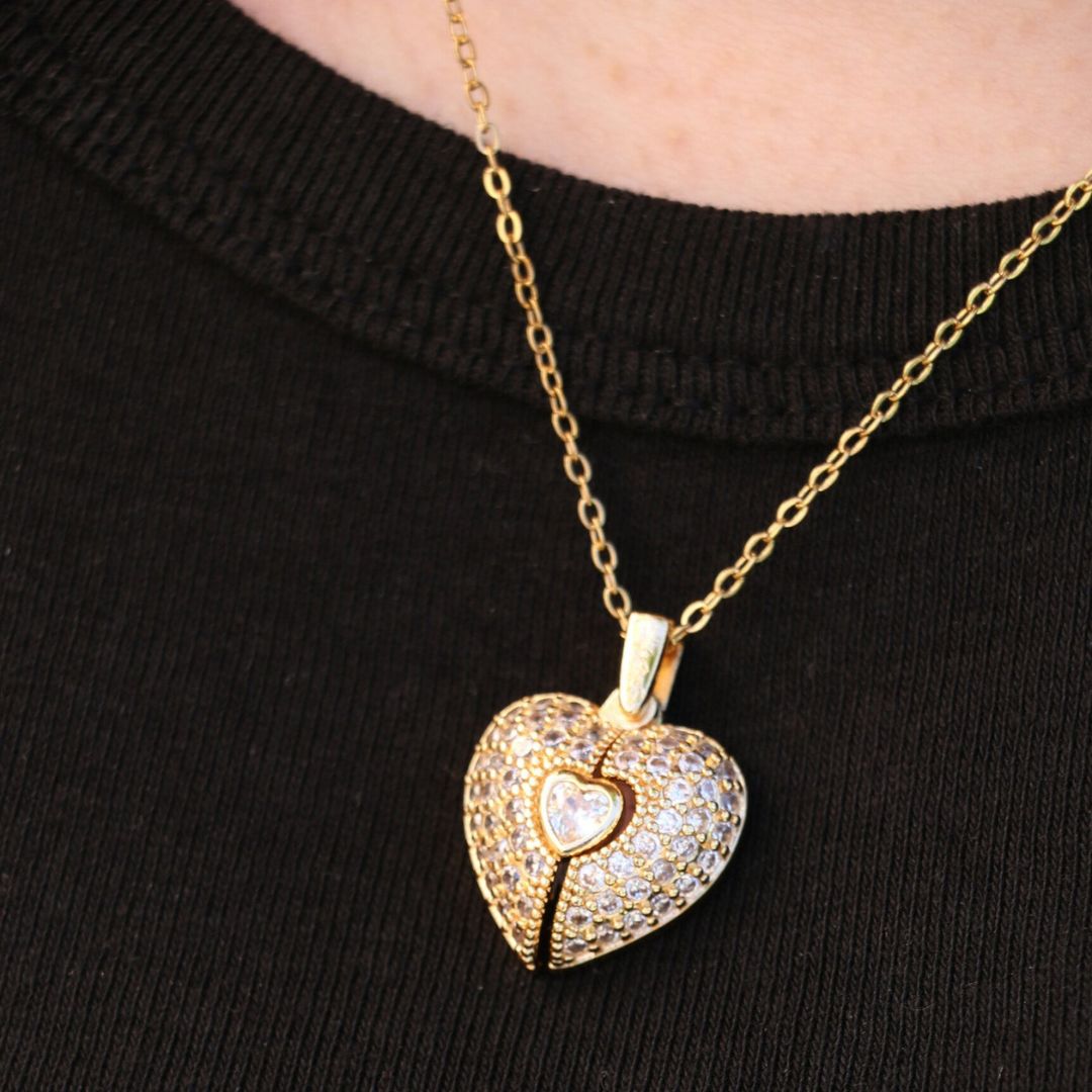 El Collar de Corazón Cofre personalizado es un collar de cadena dorada con un colgante en forma de corazón incrustado de pequeños cristales y un corazón central, que se muestra sobre un top negro de canalé.