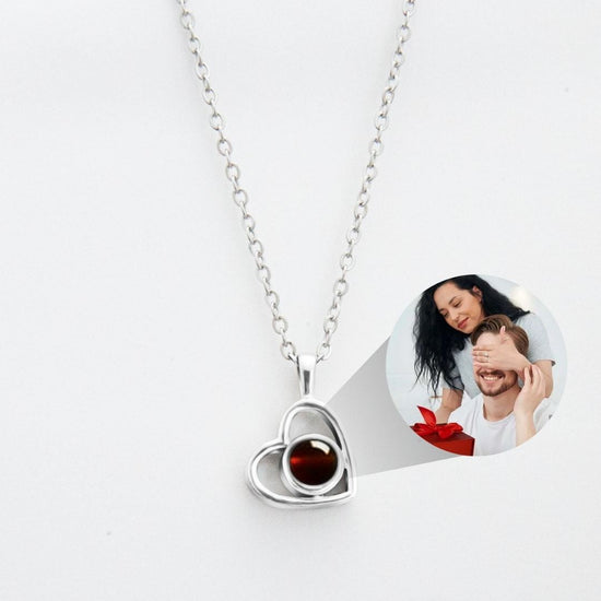 Un collar de corazón con foto, de plata y centro oscuro, cuelga de una cadena. Una imagen muestra a una mujer sorprendiendo a un hombre con una caja roja, ideal para crear recuerdos especiales con este Collar de corazón con foto.