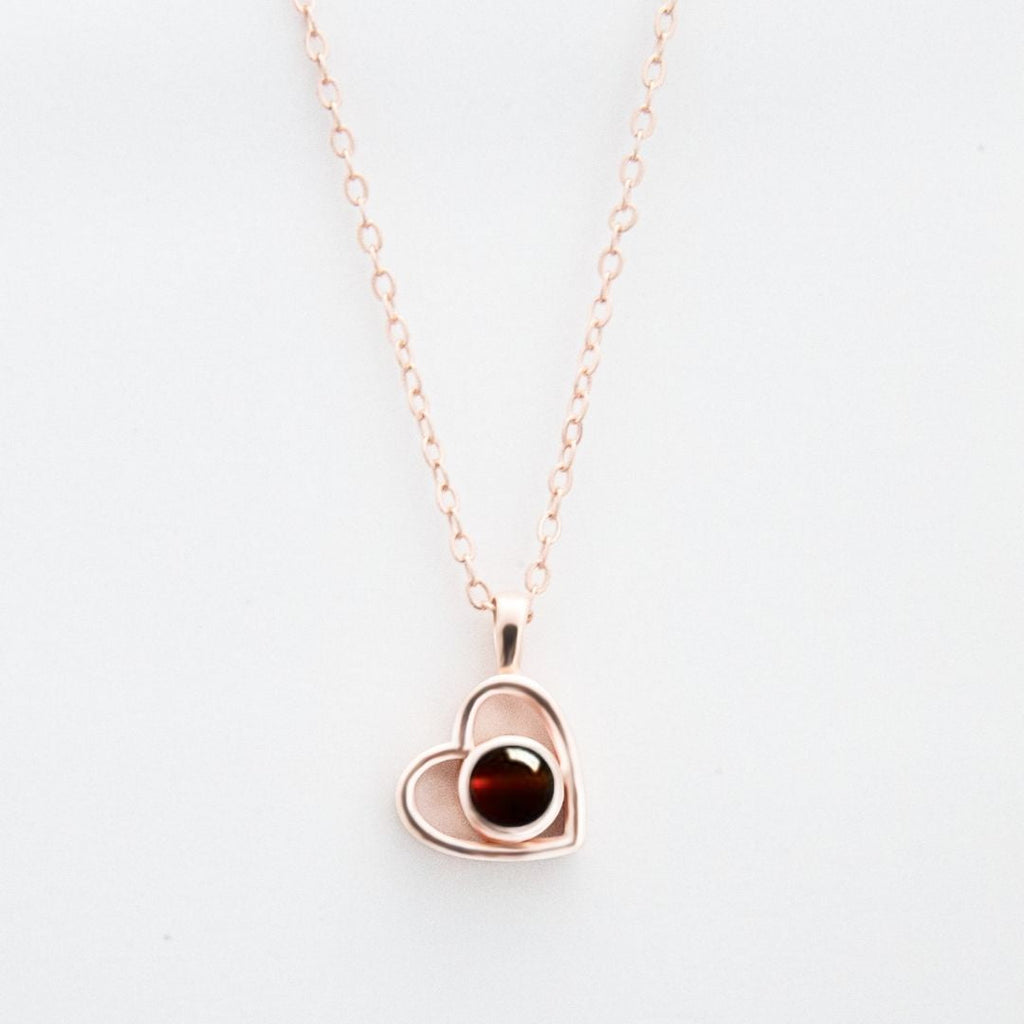 El Collar de corazón con foto es una delicada cadena en oro rosa con colgante en forma de corazón y gema roja, ideal para guardar recuerdos especiales, bellamente presentado sobre fondo blanco.
