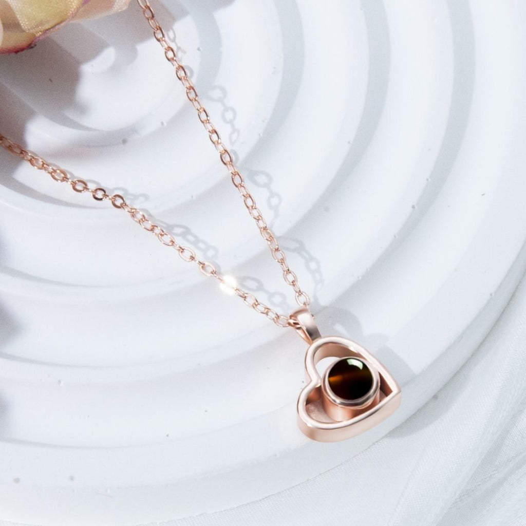 Un collar de corazón con foto en oro rosa, decorado con una piedra oscura y brillante en el centro, expuesto sobre un fondo blanco texturizado; ideal para guardar recuerdos especiales.