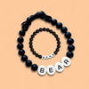 El Collar de cuentas personalizado para mascotas + Pulsera gratis se muestra con cuentas negras y grandes letras blancas BEAR sobre fondo durazno, ideal como regalo para amantes de mascotas o pulsera a juego para tu amigo peludo.