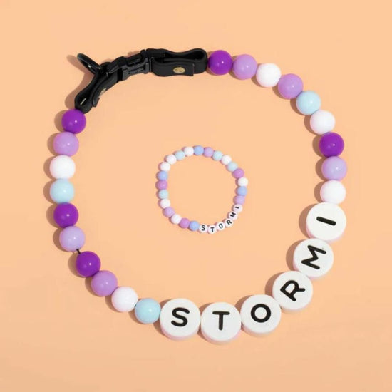 Un collar de cuentas personalizado para mascotas en tonos pastel y una pulsera gratis a juego, ambos con cuentas redondas blancas con la inscripción "STORMI", se muestran sobre un fondo melocotón, perfecto como regalo para los amantes de las mascotas.