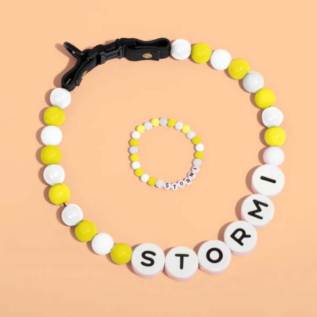El Collar de cuentas personalizado para mascotas + Pulsera gratis incluye cuentas amarillas, blancas y negras con "STORMI" en letras grandes, presentado sobre fondo melocotón-ideal como regalo para amantes de mascotas.
