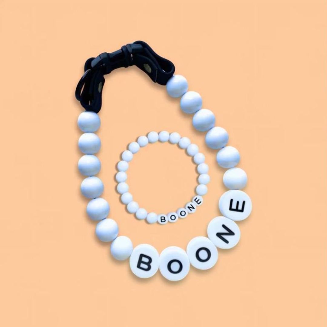 El Collar de cuentas personalizado para mascotas + Pulsera gratis con el nombre "BOONE" en cuentas blancas y negras se muestra sobre fondo durazno, siendo el regalo perfecto para amantes de mascotas.
