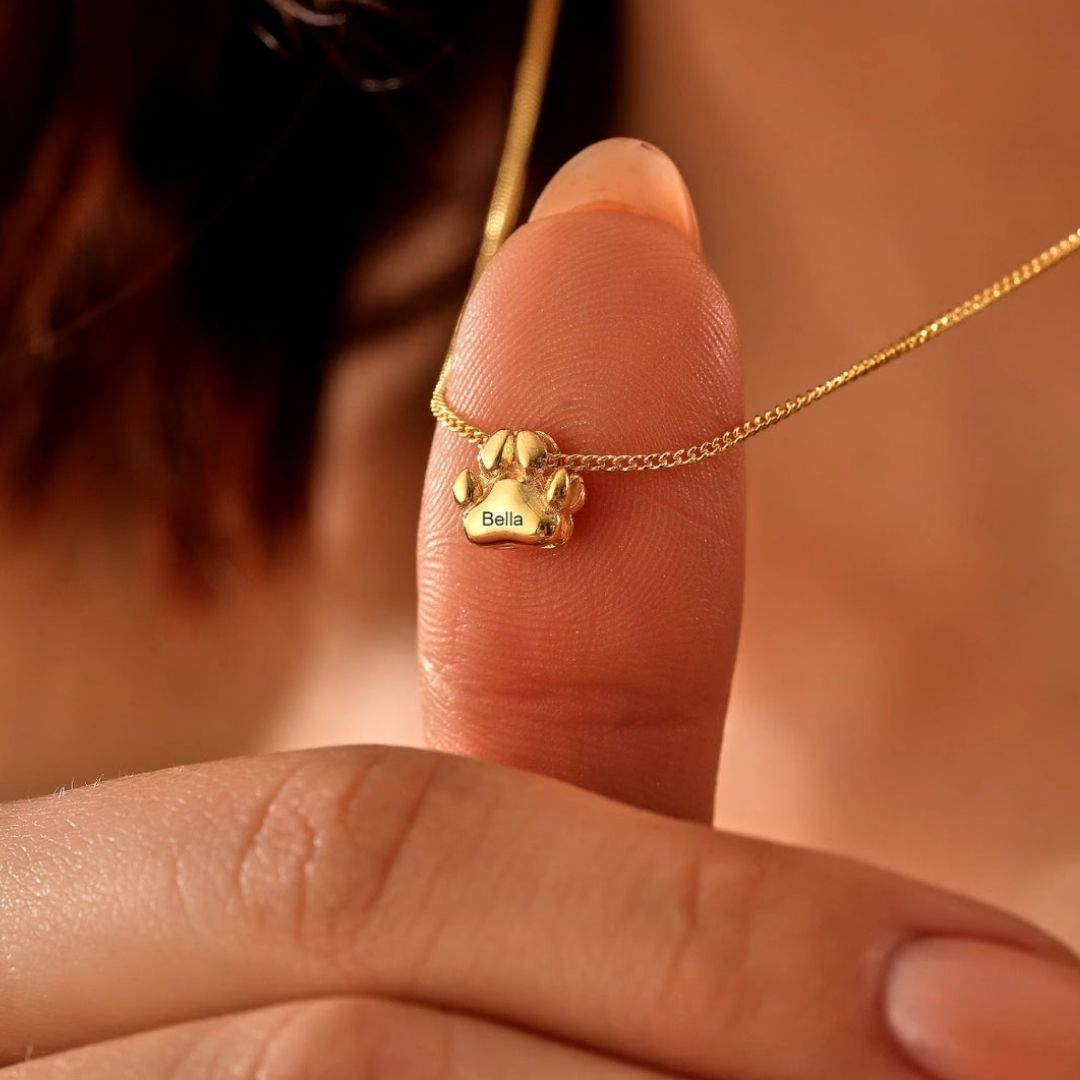 Una persona sostiene entre el pulgar y el índice un "Collar de huellitas de mascotas" personalizado con un colgante en forma de pata grabado con el nombre de Bella.