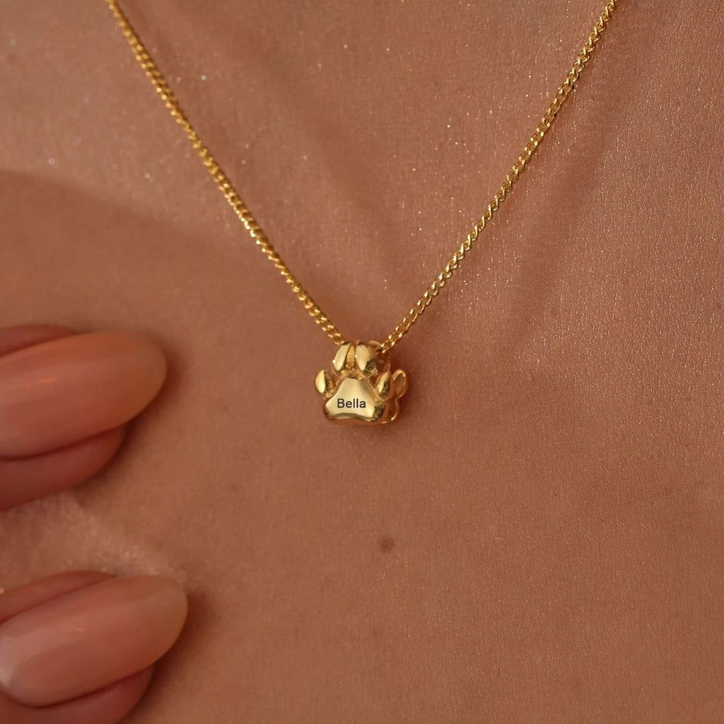 Un collar de huellitas de mascota personalizado con un colgante dorado en forma de pata grabado "Bella" cuelga de un cuello, mientras una mano con uñas desnudas toca suavemente el elegante accesorio.