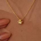 Un collar de huellitas de mascota personalizado con un colgante dorado en forma de pata grabado "Bella" cuelga de un cuello, mientras una mano con uñas desnudas toca suavemente el elegante accesorio.