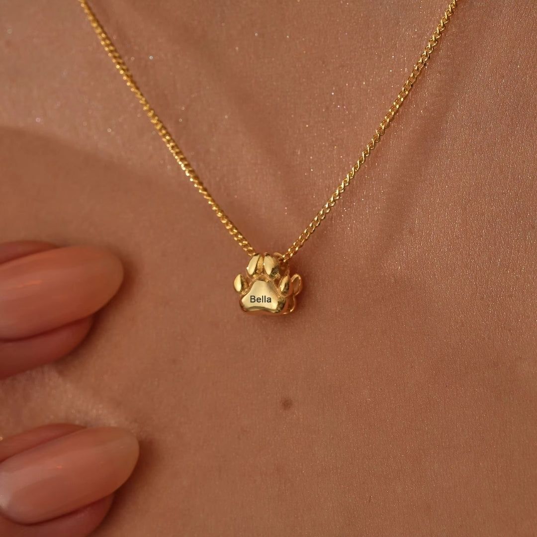 Un collar de huellitas de mascota personalizado con un colgante dorado en forma de pata grabado "Bella" cuelga de un cuello, mientras una mano con uñas desnudas toca suavemente el elegante accesorio.
