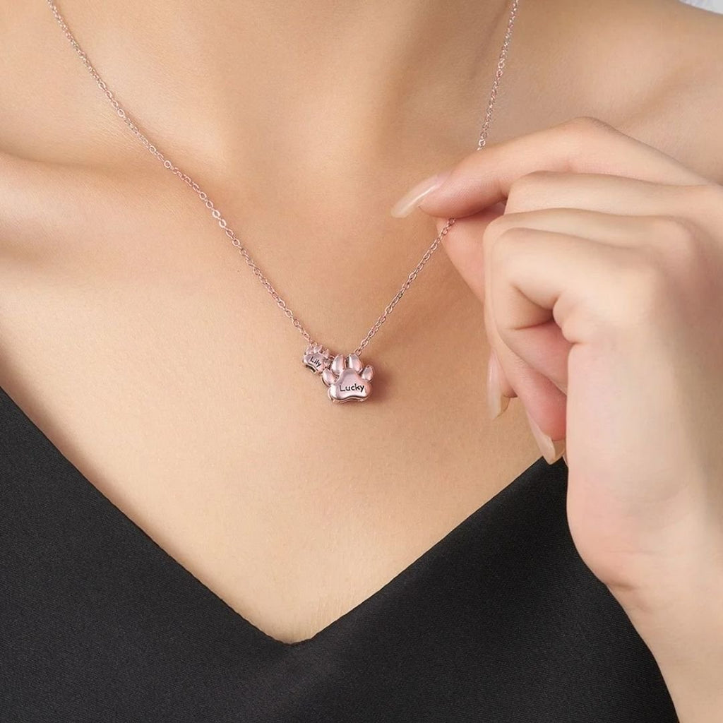 Una mujer con top negro muestra el "Collar de huellitas de mascotas personalizado", con un colgante en forma de pata grabado con "Lucky", mientras sujeta suavemente la cadena cerca de su clavícula.
