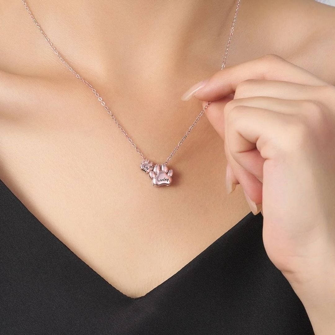 Una mujer con top negro muestra el "Collar de huellitas de mascotas personalizado", con un colgante en forma de pata grabado con "Lucky", mientras sujeta suavemente la cadena cerca de su clavícula.