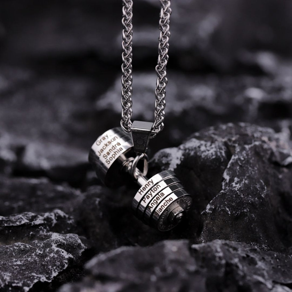 Primer plano de un collar de pesa personalizado para papá con dos colgantes cilíndricos de plata grabados con nombres, expuestos sobre una superficie de roca áspera y oscura.