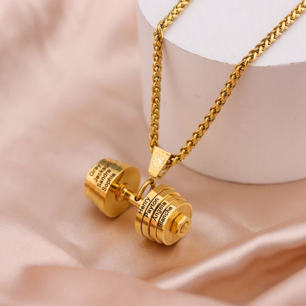 El Collar de pesa personalizado, un dije dorado en forma de mancuerna con nombres grabados en ambos lados, se exhibe sobre satén beige y un soporte blanco-regalo perfecto para papá en el Día del Padre.