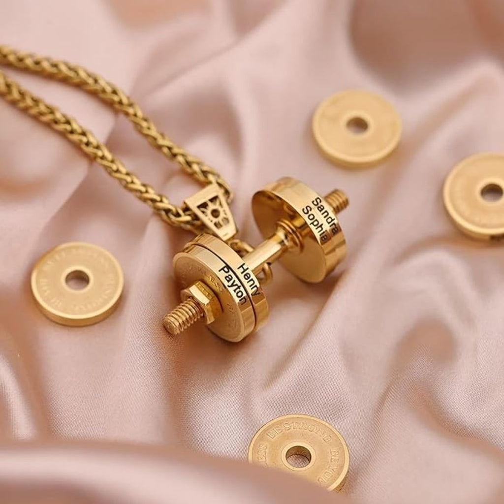El Collar de pesa personalizado, grabado con nombres, reposa sobre satén rosa junto a pequeños discos dorados. Un regalo Día del Padre perfecto para papás apasionados por el fitness.