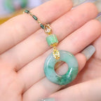 Una mano sostiene un "Collar de piedra jade esmeralda natural" que luce un dije circular tipo donut de jade, una cuenta verde pequeña y detalles dorados en un cordón verde.