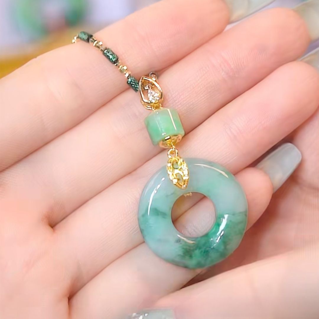 Una mano sostiene un "Collar de piedra jade esmeralda natural" que luce un dije circular tipo donut de jade, una cuenta verde pequeña y detalles dorados en un cordón verde.