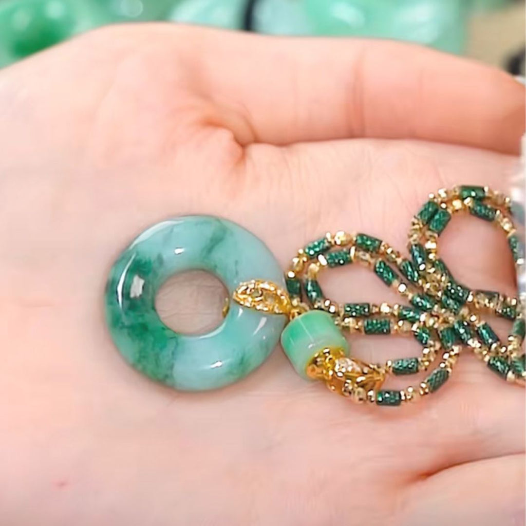 Una mano sostiene un colgante de donut de jade verde con cierre de oro, unido a un collar de cuentas verdes y doradas de piedra jade esmeralda natural. El fondo difuminado resalta la elegancia de este collar de jade natural.