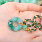 Una mano sostiene un colgante de donut de jade verde con cierre de oro, unido a un collar de cuentas verdes y doradas de piedra jade esmeralda natural. El fondo difuminado resalta la elegancia de este collar de jade natural.