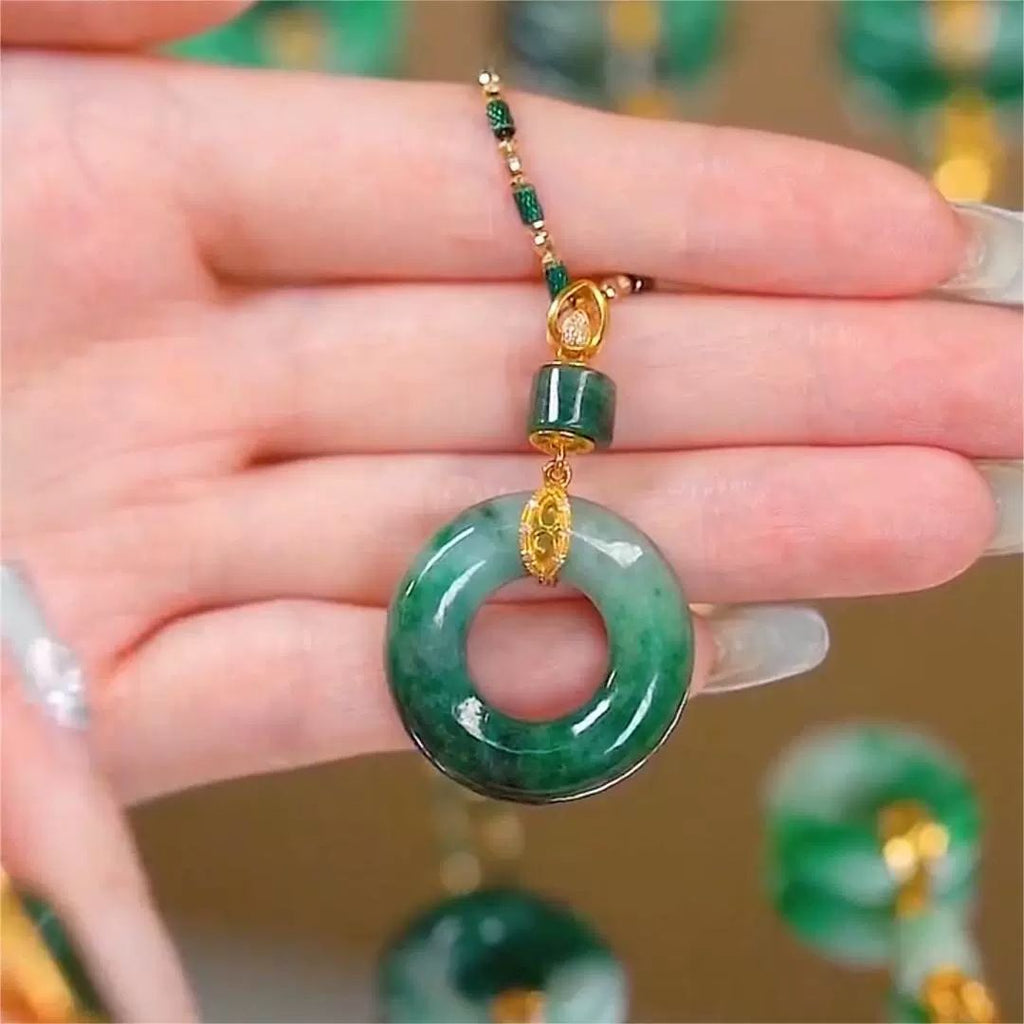 Una mano sostiene un Collar de piedra jade esmeralda natural con forma de rosquilla con jade verde brillante, intrincados detalles dorados y detalles de cuentas verdes: una llamativa pieza de joyería de jade natural.