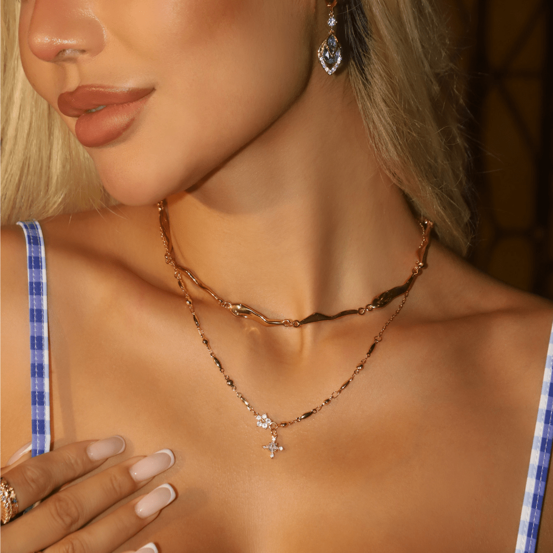 Una mujer lleva el Collar Destello de estrella -un collar gargantilla ondulado dorado- combinado con un delicado colgante en forma de cruz, pendientes en forma de gota y uñas desnudas cuidadas. Completa su look con un top de cuadros azules y blancos; su rostro es parcialmente visible.