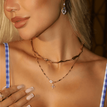 Cargar imagen en el visor de la galería, Una mujer lleva el Collar Destello de estrella -un collar gargantilla ondulado dorado- combinado con un delicado colgante en forma de cruz, pendientes en forma de gota y uñas desnudas cuidadas. Completa su look con un top de cuadros azules y blancos; su rostro es parcialmente visible.