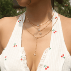 Una mujer está de pie al aire libre en medio de una exuberante vegetación, con un vestido blanco de tirantes con estampados de cerezas rojas y collares de oro en capas, incluido el collar Destello de estrella.