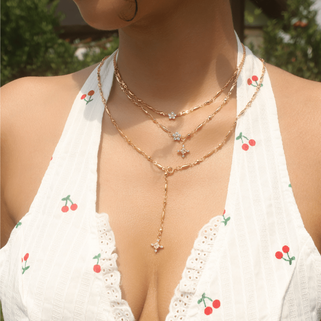 Una mujer está de pie al aire libre en medio de una exuberante vegetación, con un vestido blanco de tirantes con estampados de cerezas rojas y collares de oro en capas, incluido el collar Destello de estrella.