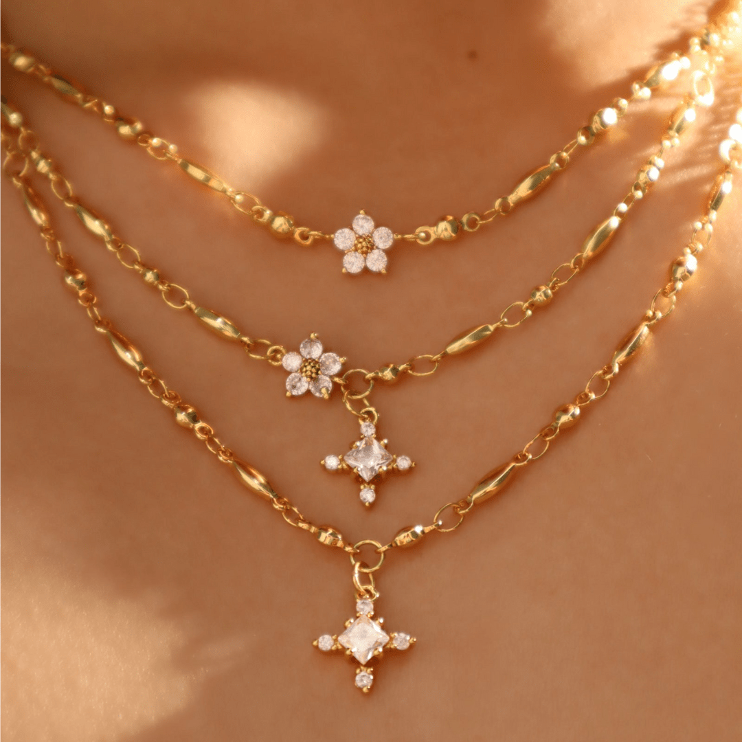 Primer plano de una persona que lleva collares de oro en capas, entre los que destaca el Collar Destello de estrella con brillantes colgantes florales y de estrella, sobre una piel cálida y suavemente iluminada.