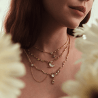 Una mujer lleva el collar Destello de estrella, un collar de oro con motivos celestes: estrellas, una luna creciente y piedras preciosas. Unas flores desenfocadas enmarcan la escena, mostrando únicamente el cuello y la parte inferior del rostro.