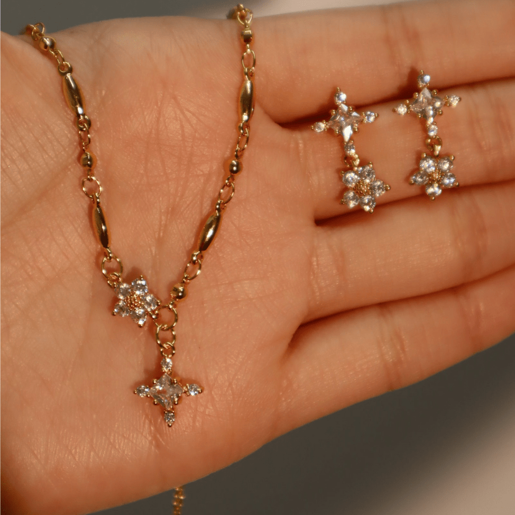 Una mano sostiene el Collar Destello de estrella, un collar de oro con un colgante en forma de estrella con brillantes cristales, junto con unos pendientes de estrella de oro a juego también adornados con cristales.