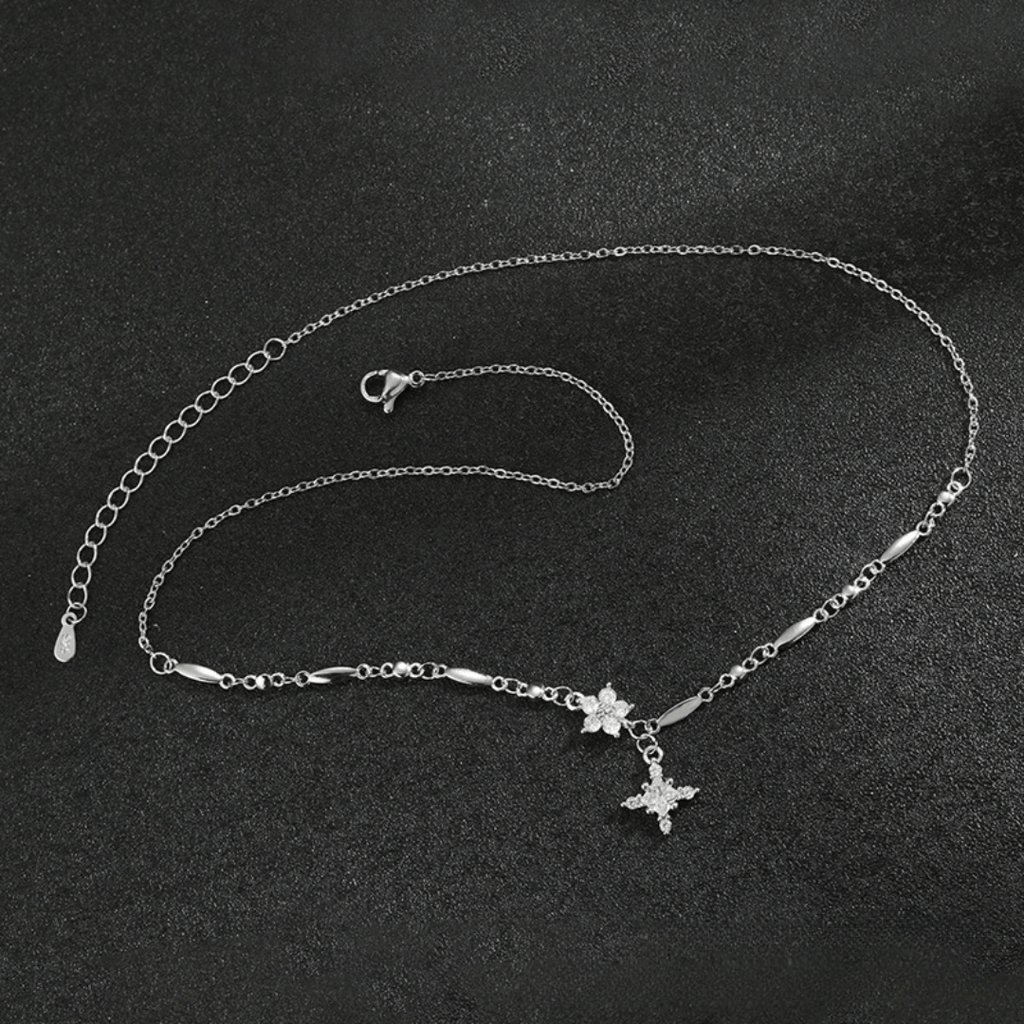 El collar Destello de estrella es un delicado collar de plata con una fina cadena, pequeños eslabones decorativos y un colgante en forma de estrella acentuado con piedras brillantes, que se muestra sobre una superficie negra texturizada.