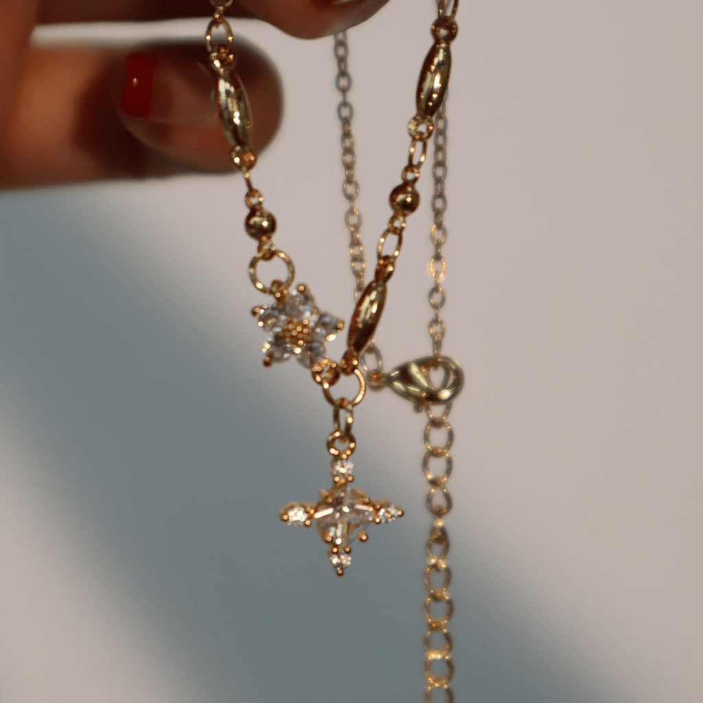 Una mano con las uñas pintadas de rojo sostiene el collar Destello de estrella, con eslabones de abalorios chapados en oro y dos colgantes en forma de estrella con brillantes piedras transparentes. El fondo suavemente difuminado resalta su elegancia.