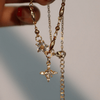 Una mano con las uñas pintadas de rojo sostiene el collar Destello de estrella, con eslabones de abalorios chapados en oro y dos colgantes en forma de estrella con brillantes piedras transparentes. El fondo suavemente difuminado resalta su elegancia.