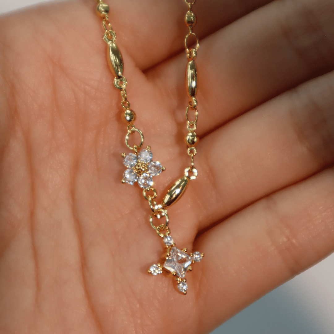 Una mano sostiene el Collar Destello de estrella, un minimalista collar chapado en oro con dos brillantes colgantes en forma de flor adornados con piedras preciosas transparentes. Su delicada cadena cuenta con pequeñas cuentas doradas y eslabones ovalados que le confieren una sutil elegancia.