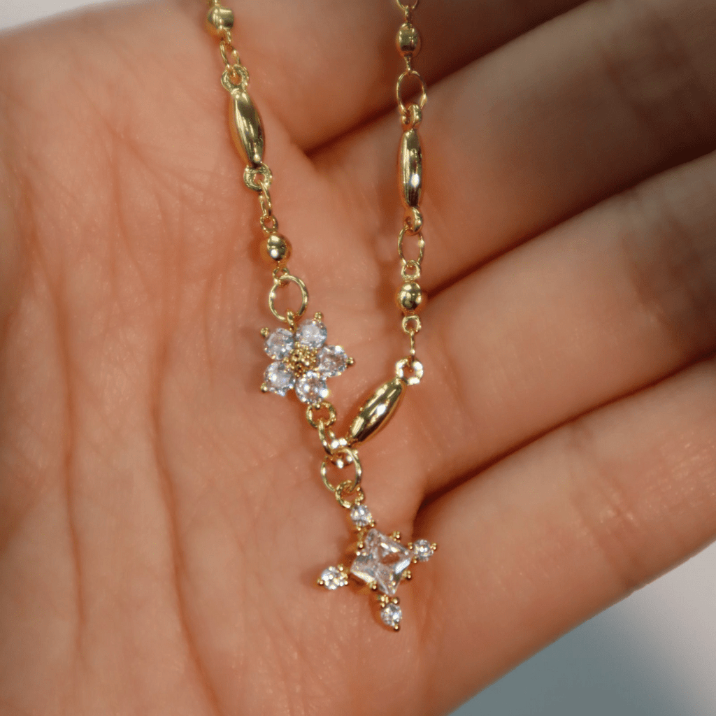 Una mano sostiene el Collar Destello de estrella, un minimalista collar chapado en oro con dos brillantes colgantes en forma de flor adornados con piedras preciosas transparentes. Su delicada cadena cuenta con pequeñas cuentas doradas y eslabones ovalados que le confieren una sutil elegancia.