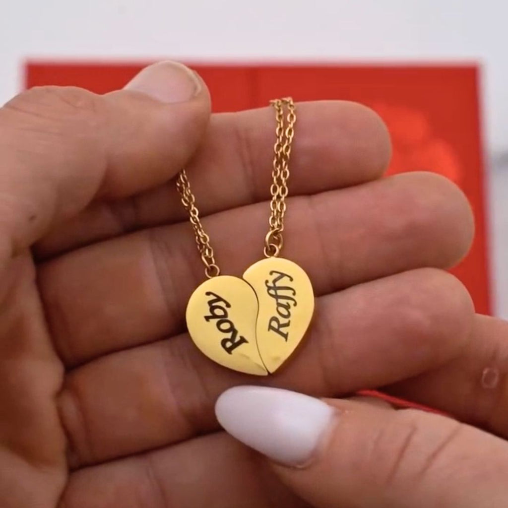 Una mano sostiene un collar Dúo medio corazón en oro, cada mitad en una cadena con "Raffy" y "Ruby" grabados-un regalo ideal para parejas. Se aprecia un toque de fondo rojo.