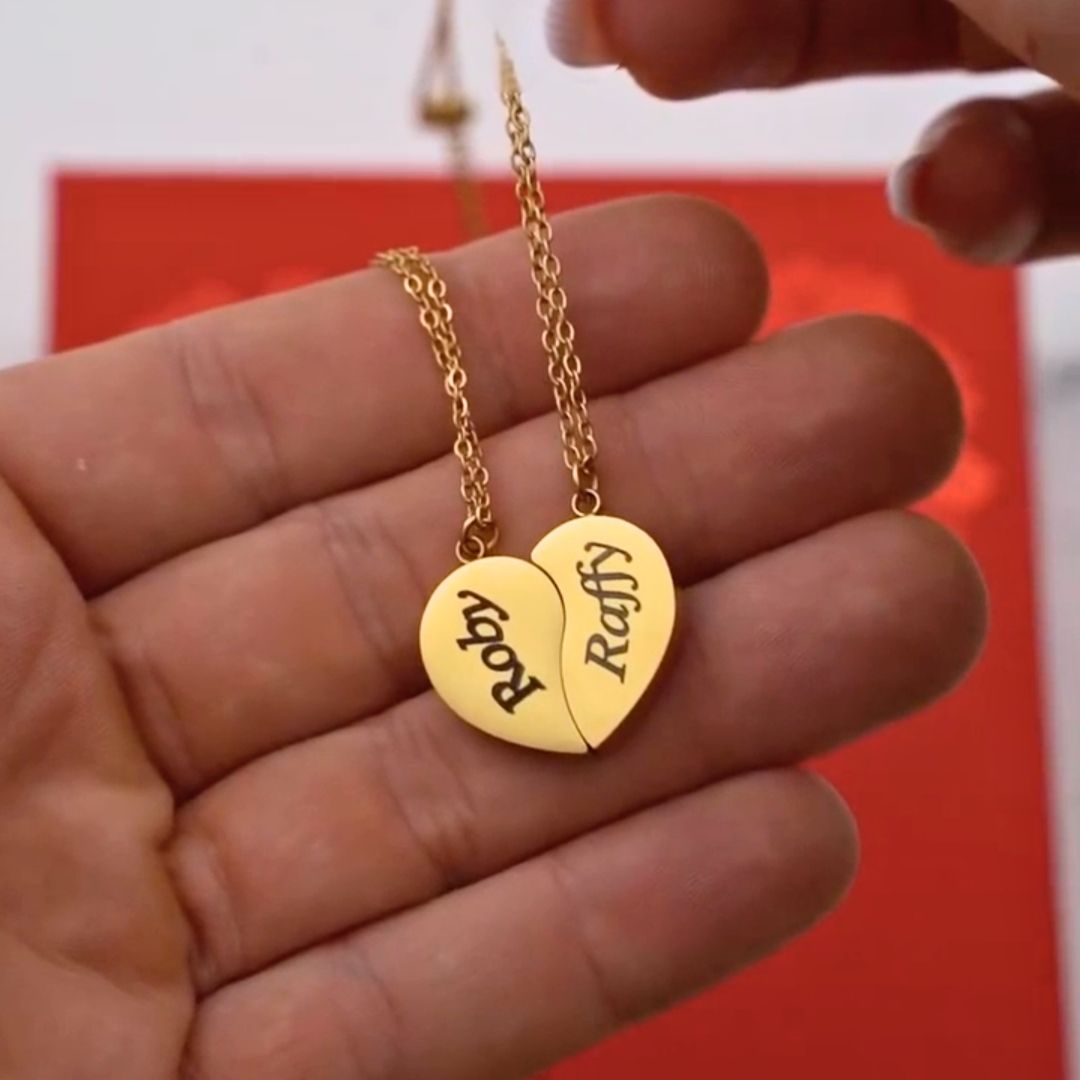Unas manos sostienen el Collar Dúo medio corazón dorado, dividido en dos cadenas: una dice Rafy y la otra Roby. Sobre fondo rojo, este collar es el regalo ideal para parejas.