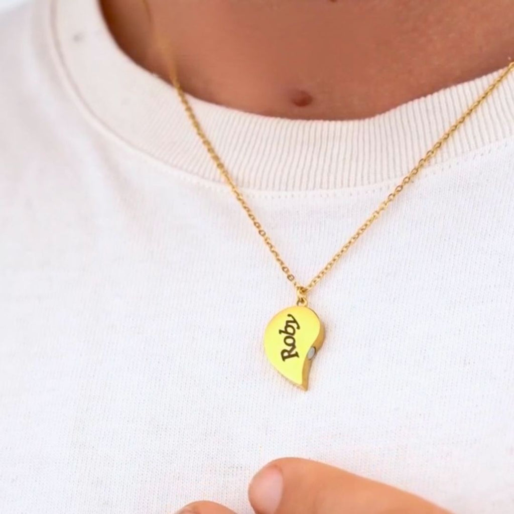 Alguien lleva el collar Dúo medio corazón de oro, grabado con "Roby" en el colgante de medio corazón, sobre una camisa blanca. Una mano toca suavemente cerca, destacando este perfecto regalo para parejas.