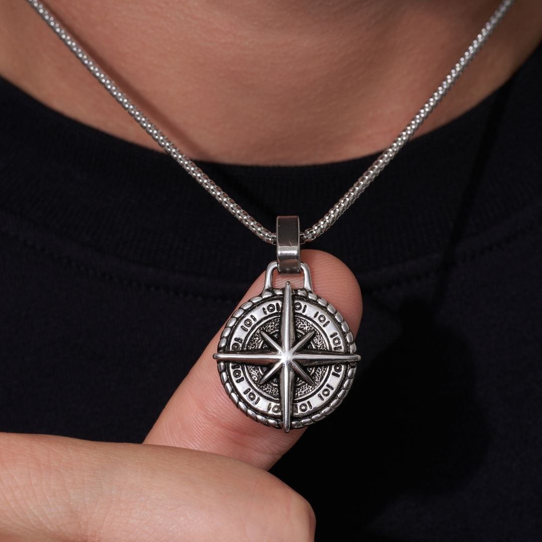 Una persona sostiene cerca de su cuello el Collar Estrella del Norte - Para mi hijo (con nota dedicada), un colgante plateado con diseño de rosa de los vientos. Lleva una prenda oscura, haciendo del collar el regalo perfecto para un hijo.
