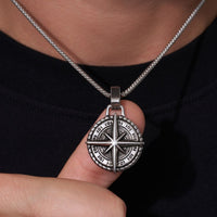 Una persona sostiene cerca de su cuello el Collar Estrella del Norte - Para mi hijo (con nota dedicada), un colgante plateado con diseño de rosa de los vientos. Lleva una prenda oscura, haciendo del collar el regalo perfecto para un hijo.