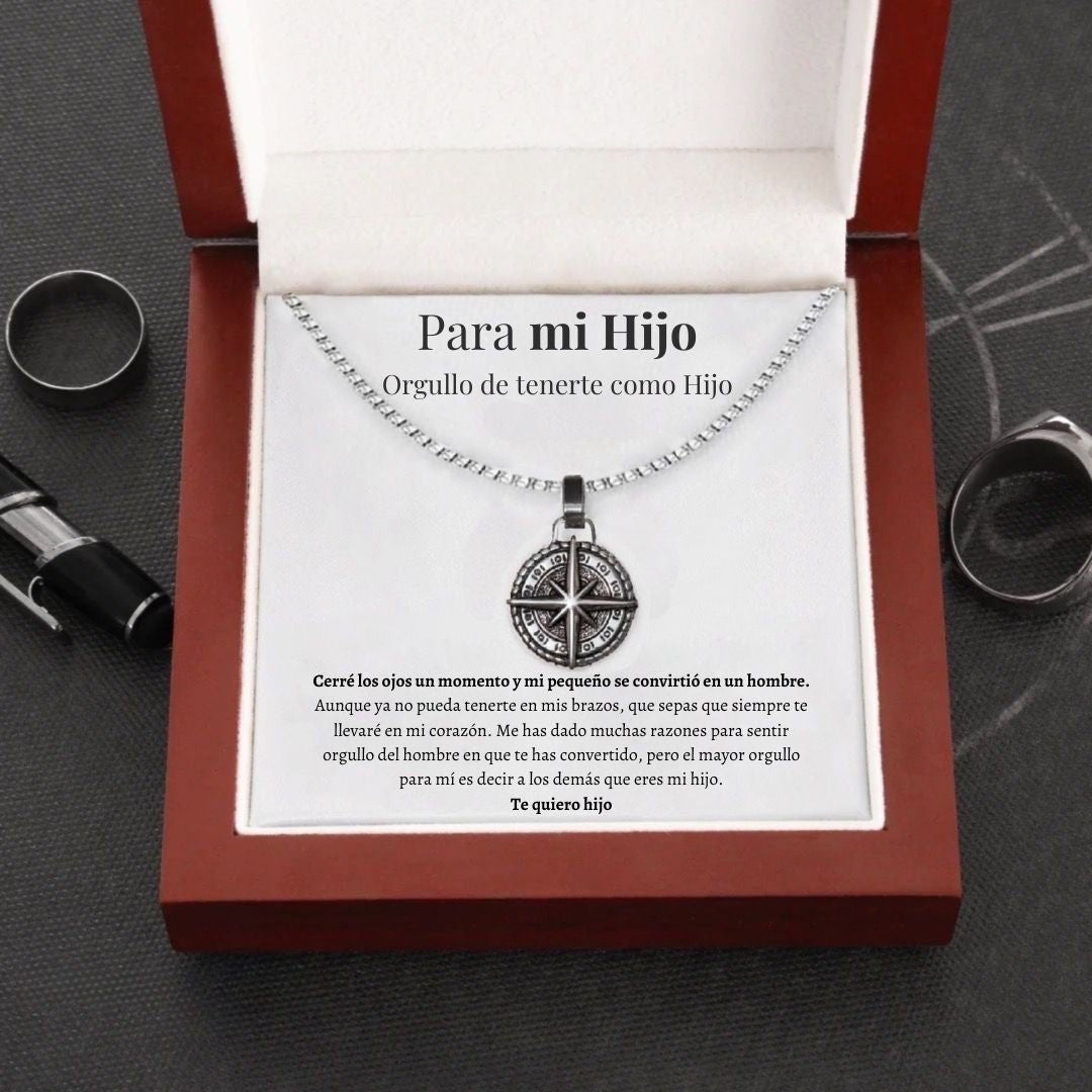 El collar Estrella del Norte - Para mi hijo (con nota dedicada) se presenta en una caja roja abierta con una cariñosa nota en español para un hijo, que expresa orgullo y amor. Rodeado de anillos y un bolígrafo sobre una superficie oscura, es un regalo lleno de significado.