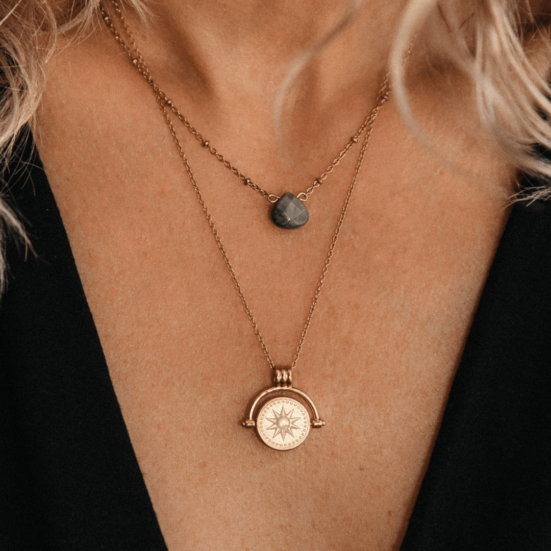 Primer plano de una persona de pelo rubio ondulado con un top negro que lleva dos collares de oro: el collar giratorio Sol y Luna "Hatha" y otro con un colgante de piedra gris.