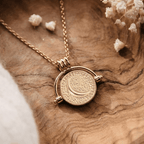 Un collar de oro, Collar giratorio Sol y Luna "Hatha", con un colgante redondo de textura que representa una luna creciente, descansa sobre madera rodeada de delicadas flores blancas secas, captando su belleza celestial.